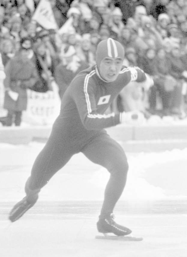 Ein Schwarz-Weiß-Foto eines Eisschnellläufers in Aktion auf dem Eis, der einen Helm und Eislaufschuhe trägt, mit ein paar Menschen im Hintergrund, die Fahnen halten.