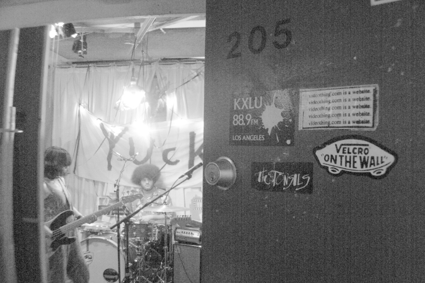 Ein Schwarz-Weiß-Bild eines Mannes, der vor Trommeln sitzt, einer Frau, die Gitarre spielt, und einem Banner mit der Aufschrift 'YUCK' im Hintergrund, mit einer sichtbaren Tür.