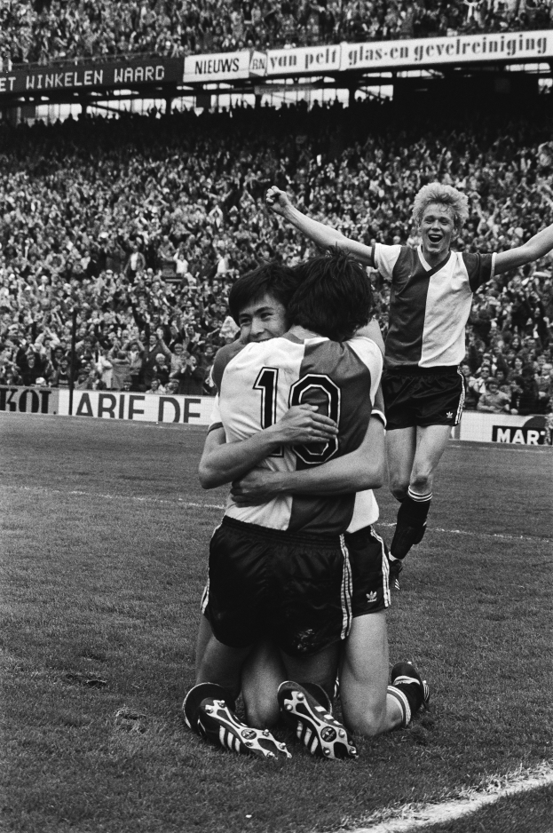 Drei Fußballspieler feiern ein Tor auf dem Boden, umgeben von einer jubelnden Menge in einem schwarz-weißen Stadion-Szenario.