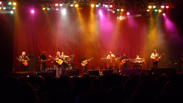 Eine Theateraufführung mit ein paar Gitarren spielenden Menschen, einem Vorhang im Hintergrund und einer kleinen Gruppe von Menschen am Bildrand.