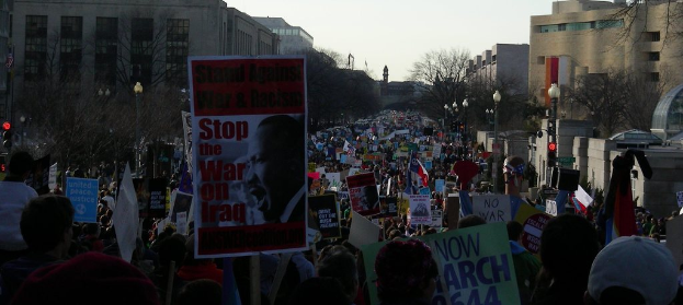 Eine Gruppe von Menschen demonstriert auf einer Straße, hält Plakate hoch, mit Gebäuden, Bäumen, Laternenpfählen und dem Himmel im Hintergrund.
