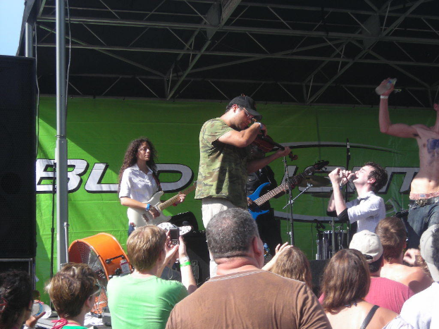 Eine Gruppe Musiker auf der Bühne mit verschiedenen Instrumenten, ein großer schwarzer Lautsprecher links und ein Publikum vor einem Podium mit einem grünen Banner im Hintergrund.