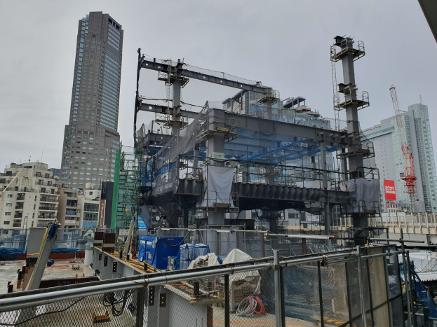 Großbaustelle in einer Stadt mit teilweise errichteter Kraftwerkumrandung von einem Metallzaun, Fahrzeugen, Strommasten, einem Banner und einem bewölkten Himmel.