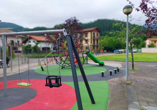 Spielplatz mit Schaukeln, grüner Rutsche, Pfosten, Lichtern, Bänken, Gras, Bäumen, Gebäuden, Fahrzeugen, Mülltonnen, Hängen und einem klaren blauen Himmel.