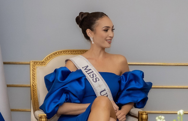 Eine Frau in einem blauen Kleid trägt eine 'Miss Universe 2019'-Schärpe und sitzt lächelnd auf einem Stuhl umgeben von Blumen mit einer Wand im Hintergrund.
