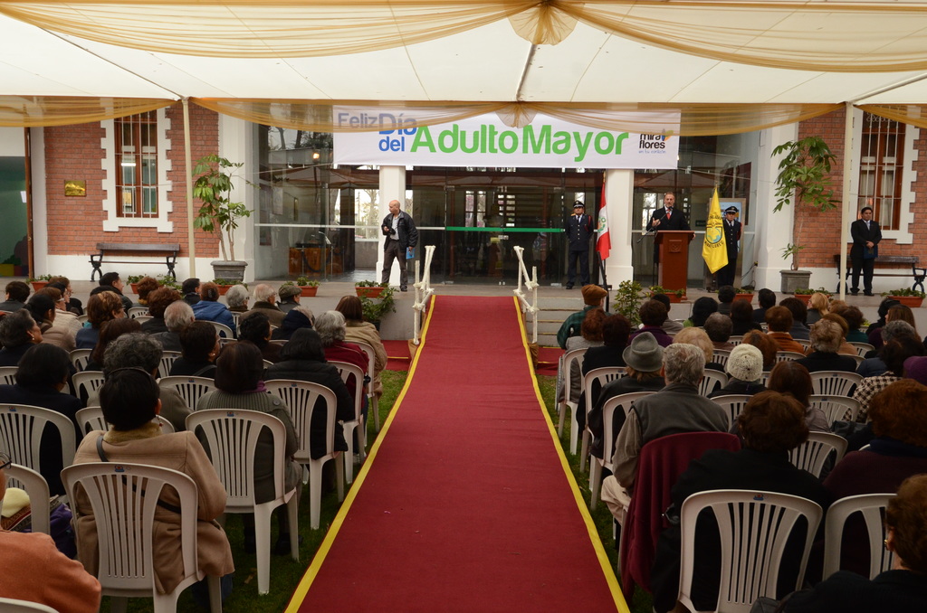 Ein Mann steht vor einem Gebäude mit einem Bogen, auf dem 'Adulto Mayor' steht, während viele Menschen im Hintergrund auf Stühlen sitzen.