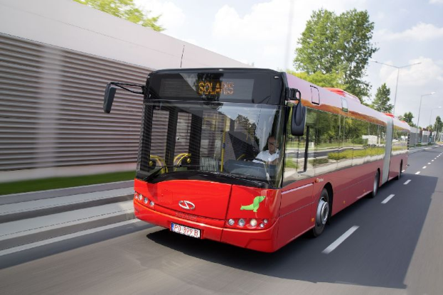 Ein roter Bus fährt auf einer Straße an einem Gebäude vorbei, in dem sich eine Person befindet, während sich auf der linken Seite eine Wand befindet und im Hintergrund Bäume, Laternenpfähle und ein bewölkter Himmel befinden.