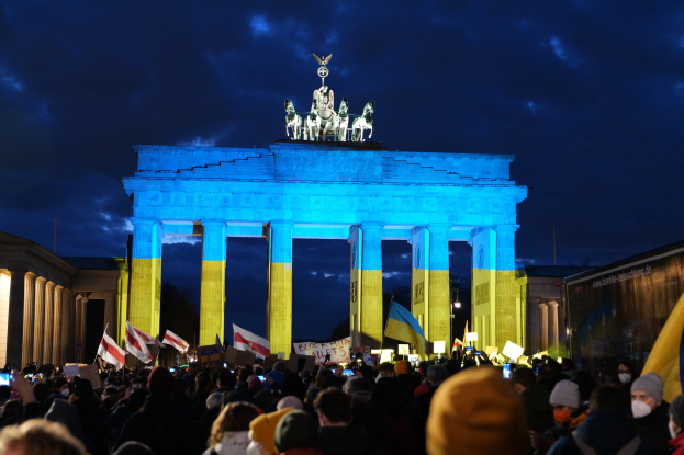 Eine Menschenmenge mit Fahnen und Plakaten steht vor dem Brandenburger Tor in Berlin, Deutschland, mit einer Fahne auf der rechten Seite und bewölktem Himmel.