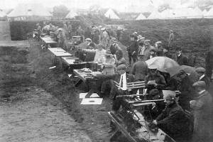 Eine Gruppe von Männern in einem schwarz-weißen Setting steht um einen Tisch mit Gewehren, einige halten Schirme, mit Zelten, Bäumen und einem klaren Himmel im Hintergrund, was auf einen Schießwettbewerb hindeutet.