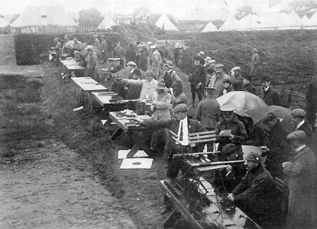Eine Gruppe von Männern in einem schwarz-weißen Setting steht um einen Tisch mit Gewehren, einige halten Schirme, mit Zelten, Bäumen und einem klaren Himmel im Hintergrund, was auf einen Schießwettbewerb hindeutet.