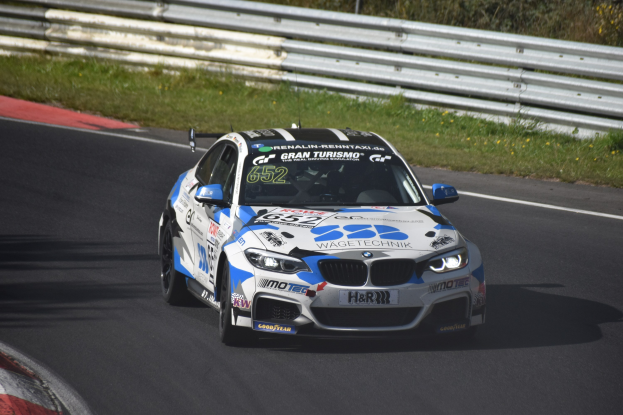 Ein BMW M4 DTM-Rennwagen mit Text und Zahlen auf seiner Karosserie, der auf einer Rennstrecke fährt, umgeben von Gras, einem Zaun und Pflanzen, wobei ein Rad sichtbar ist.