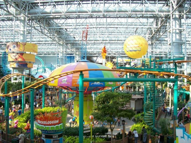 Ein großer Indoor-Wasserpark mit einer Achterbahn, umgeben von Menschen, Bäumen, Pflanzen und beleuchteten Deckenstangen und -lichtern.