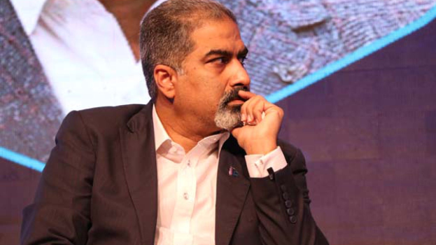 Ein Mann in Anzug und Krawatte sitzt auf einer Bühne, in Gedanken versunken, einem Bildschirm gegenüber.