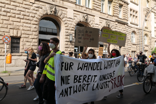 Eine Gruppe von Menschen marschiert auf einer Straße in Berlin, hält Schilder und Banner hoch und fährt mit Fahrrädern an einem Gebäude mit Bögen, Säulen, Skulpturen und Bäumen vorbei.