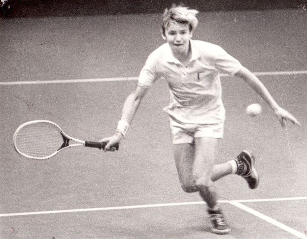 Junger Mann in weißer Tennisbekleidung schlägt auf einem Tennisplatz mit einem Schläger auf einen Ball ein.