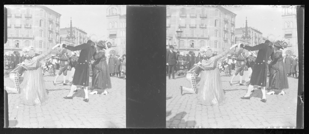Ein Schwarz-Weiß-Foto einer Gruppe von Menschen, die auf der Straße tanzen, während eines Karnevals-Umzugs, mit Kostümen, umgeben von Gebäuden, Straßenlaternen und einer klaren Himmel.