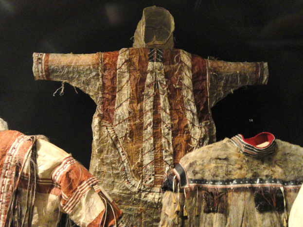 Eine Gruppe von Schaufensterpuppen in einer Museumsausstellung, die traditionelle Kleidung aus verschiedenen Materialien wie Fell und Stoff tragen, vor einem dunklen Hintergrund.