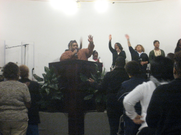 Ein Mann steht neben einem Podium auf einer Bühne und hält ein Mikrofon in der Hand, während andere Menschen hinter ihm und auf dem Boden stehen.