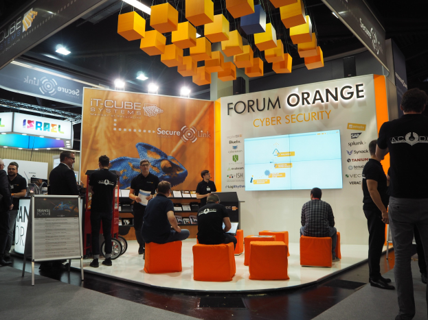 Eine Gruppe von Menschen an einem IT-Cube Systems Forum Orange Cyber Security Stand auf einer Messe, mit Stühlen, einem Display-Bildschirm, Regalen, Schildern, Deckenleuchten und einer Tafel mit Text.