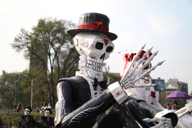 Ein Día de los Muertos-Umzug in Mexiko-Stadt mit Menschen in Kostümen, die Schirme halten und Skelette, Gebäude, Bäume und Laternenmasten im Hintergrund sowie Text am unteren Bildrand.