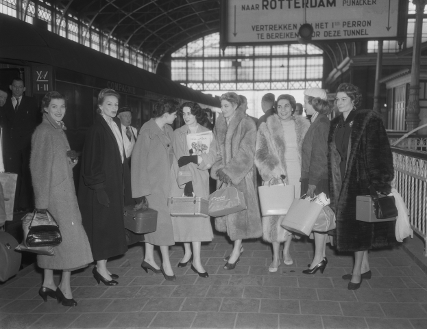 Schwarz-weißes Bild von Frauen, die an einem Bahnhof stehen und Taschen tragen, neben einem stehenden Zug und Geländern mit einer Tafel an der Decke.