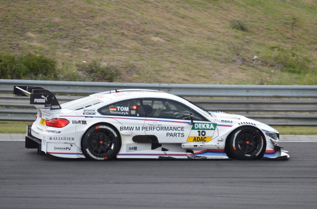 BMW M4 DTM race car on a track with sponsor text, surrounded by green grass and a railing in the background.