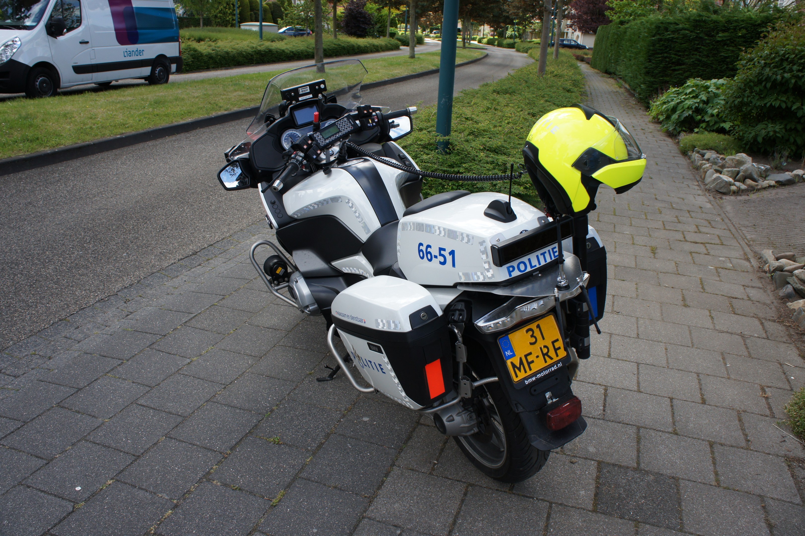 Ein Polizeimotorrad mit einem Helm auf dem Sitz, das am Straßenrand mit Bäumen, Strommasten, Gebäuden und anderen Fahrzeugen im Hintergrund geparkt ist.