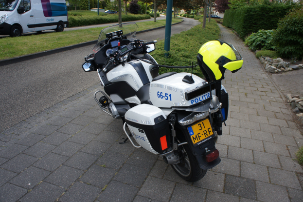 Ein Polizeimotorrad mit einem Helm auf dem Sitz, das am Straßenrand mit Bäumen, Strommasten, Gebäuden und anderen Fahrzeugen im Hintergrund geparkt ist.