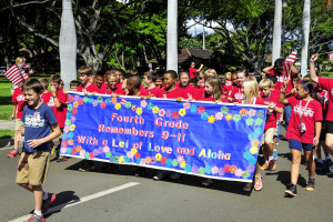Gruppe von Kindern in roten Hemden auf einer Straße, die eine Fahne und Flaggen tragen, mit Bäumen und Häusern im Hintergrund.