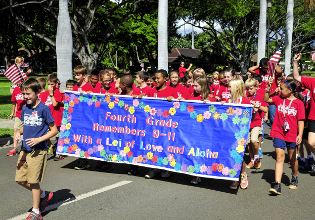Gruppe von Kindern in roten Hemden auf einer Straße, die eine Fahne und Flaggen tragen, mit Bäumen und Häusern im Hintergrund.