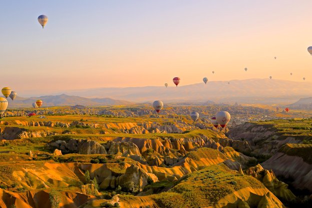Heißluftballons fliegen über das Tal von Cappadocia, Türkei, mit Fallschirmen am Himmel und Hügeln und Gras am Boden darunter.