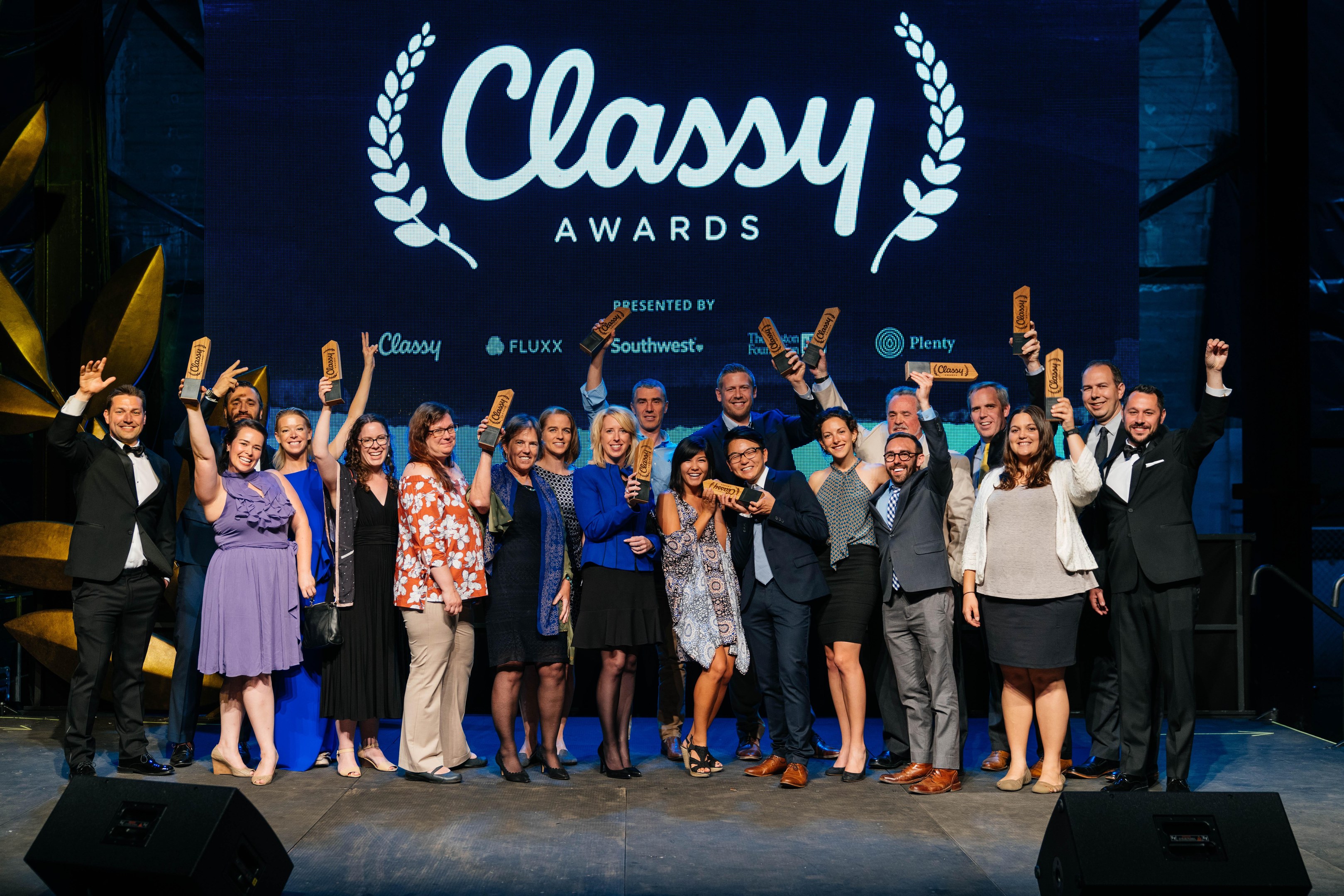 Gruppe von Menschen auf einer Bühne mit Auszeichnungen, mit Lautsprechern darunter und einem Bildschirm im Hintergrund mit "Classy Awards 2019"