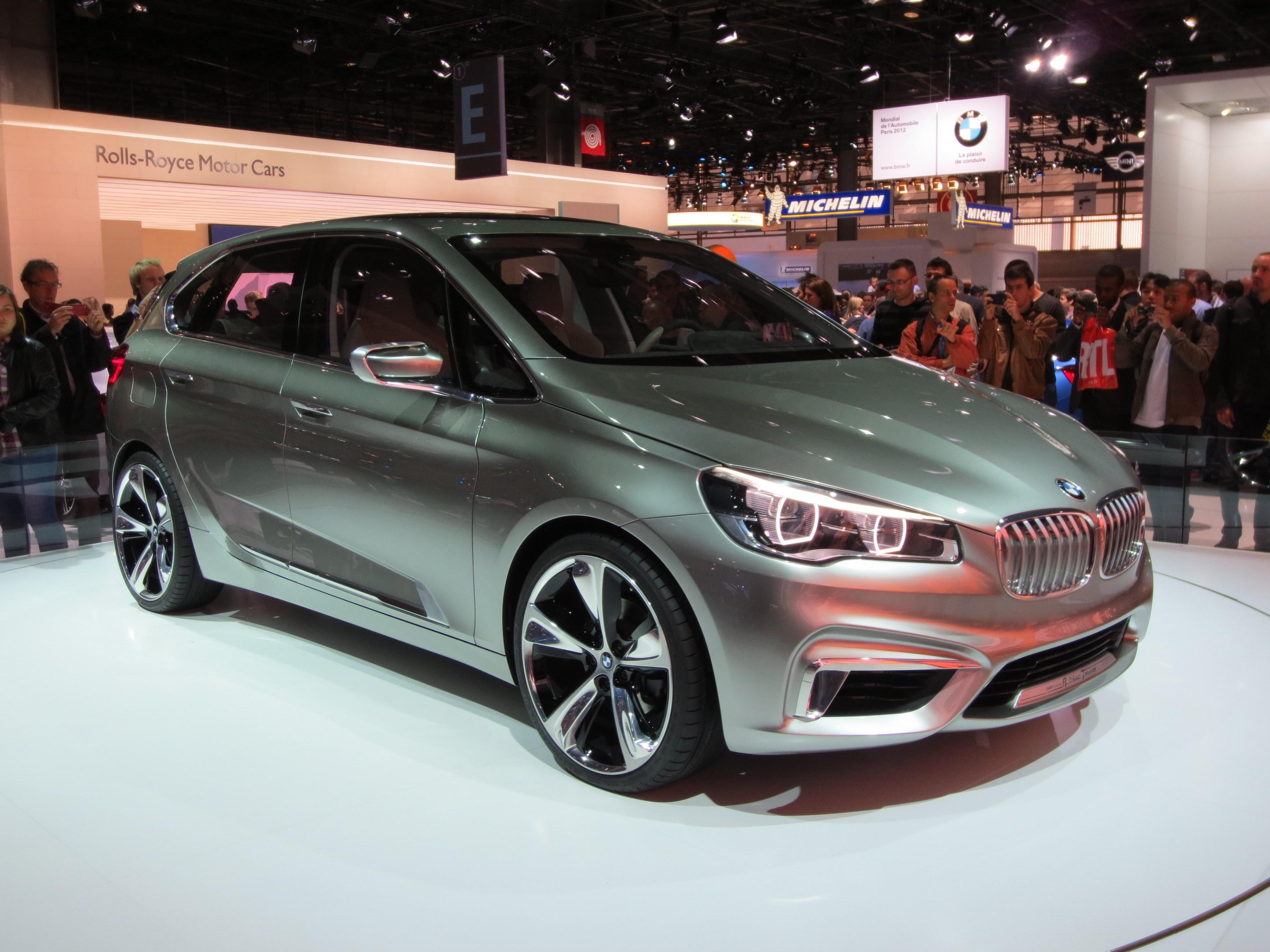 BMW 2er Active Tourer Concept auf der IAA in Frankfurt zu sehen, umgeben von Menschen, die es fotografieren, unter Deckenbeleuchtung mit Texttafeln im Hintergrund.