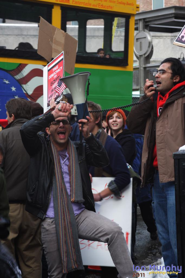 Eine Gruppe von Menschen mit Schildern, Rednern und Mikrofonen, mit einem Bus und Gebäuden im Hintergrund.
