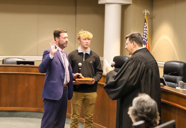 Ein Mann im Anzug und Krawatte steht neben einem Richter in einer schwarzen Robe in einem Gerichtssaal, hält ein Mikrofon und ein Buch in der Hand, mit Stühlen, Tischen, einer Flagge, einer Säule und einer Wand im Hintergrund und einer Person, die in einem Stuhl sitzt.