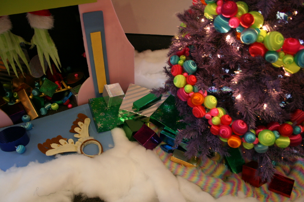 Drinnen steht rechts ein geschmückter Weihnachtsbaum mit Lichterkette und Schmuck, in der Mitte Geschenkboxen und Dekorationen, in der linken oberen Ecke eine Hütte mit Dekorationen und unten Wolle und Watte.