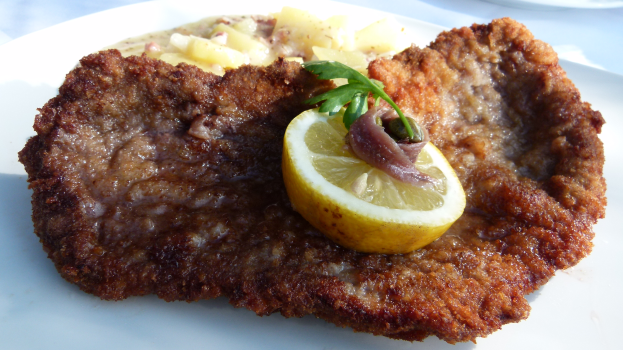 Ein weißer Teller mit einem panierten Schweinefleischstück, Kartoffeln und einer Zitronenscheibe auf einem Tisch.