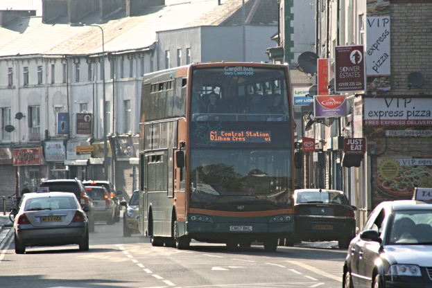 Eine Straße mit Autos und einem Bus vor Gebäuden mit Wänden, Fenstern, Tellern und Dächern, mit Plakaten und Bannern an den Wänden und einem Pfahl mit einer Straßenlaterne.