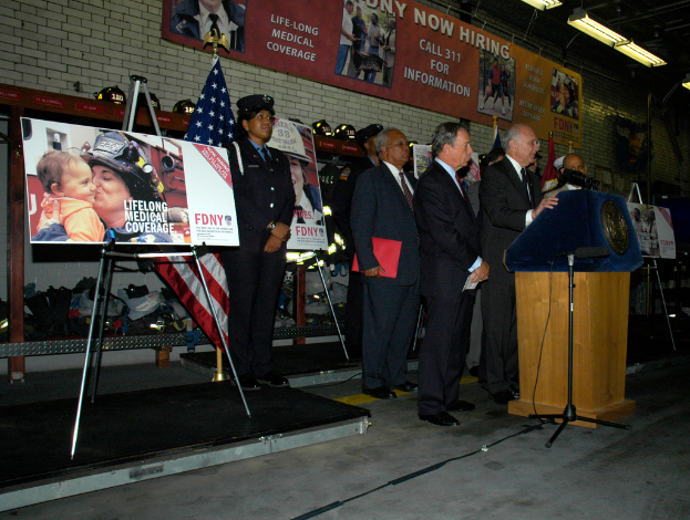 Eine Gruppe von Menschen, die in der Nähe eines Podiums mit Mikrofonen stehen, umgeben von Schildern auf Ständern, Flaggen und Schildern an der Wand, alles unter hellen Lichtern.