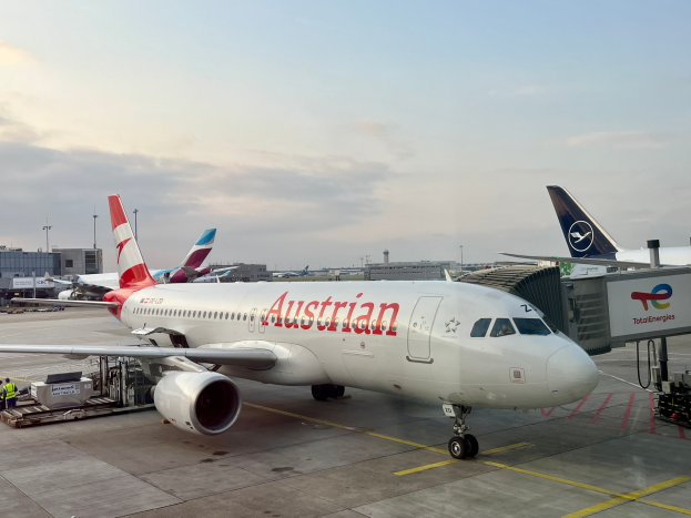 Ein Austrian-Airlines-Airbus-A330-300 auf dem Rollfeld des Frankfurter Flughafens, umgeben von Menschen, Fahrzeugen und anderen Gegenständen, mit Gebäuden, Masten und einem klaren blauen Himmel im Hintergrund.