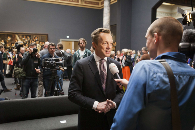 Ein Mann in Anzug und Krawatte hält ein Mikrofon und wird von einem Reporter vor einem Publikum mit Kameras interviewt, ein Sofa im Vordergrund, ein gerahmtes Foto an der Wand und Beleuchtungsausrüstung.