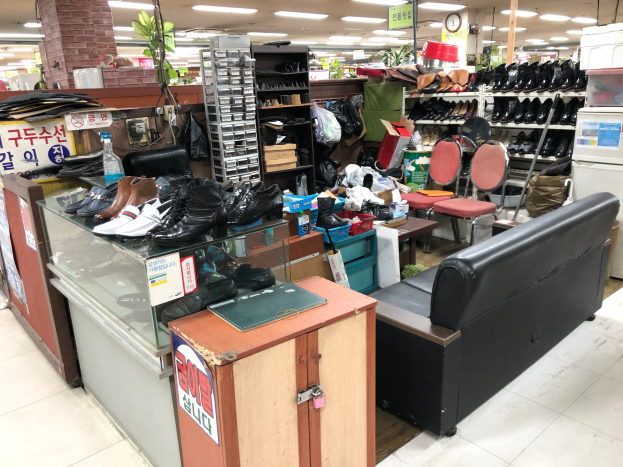 Ein Geschäft in Seoul, Südkorea, voller verschiedener Arten von Schuhen, Möbeln einschließlich einer Couch, Stühlen, Tischen und einem Kühlschrank, Schuhständern und anderen Gegenständen, einer Uhr an der Wand und Lampen an der Decke.