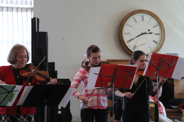 Drei Frauen auf der Bühne mit Violinen stehend und spielend, mit Notenpulten und Noten davor, während ein Zuschauer im Vordergrund sitzt; eine Uhr hängt an der weißen Wand im Hintergrund, und ein weißer Vorhang ist links sichtbar.