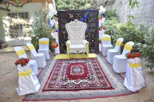 Eine Hochzeitszeremonie in einem Garten mit weißen Stühlen, einem roten Teppich, Blumenbouquets, Dekorationsgegenständen, Ballons, Pflanzen, Bäumen, einem Gebäude mit Fenstern und einer Tür und einer Wand auf der rechten Seite.