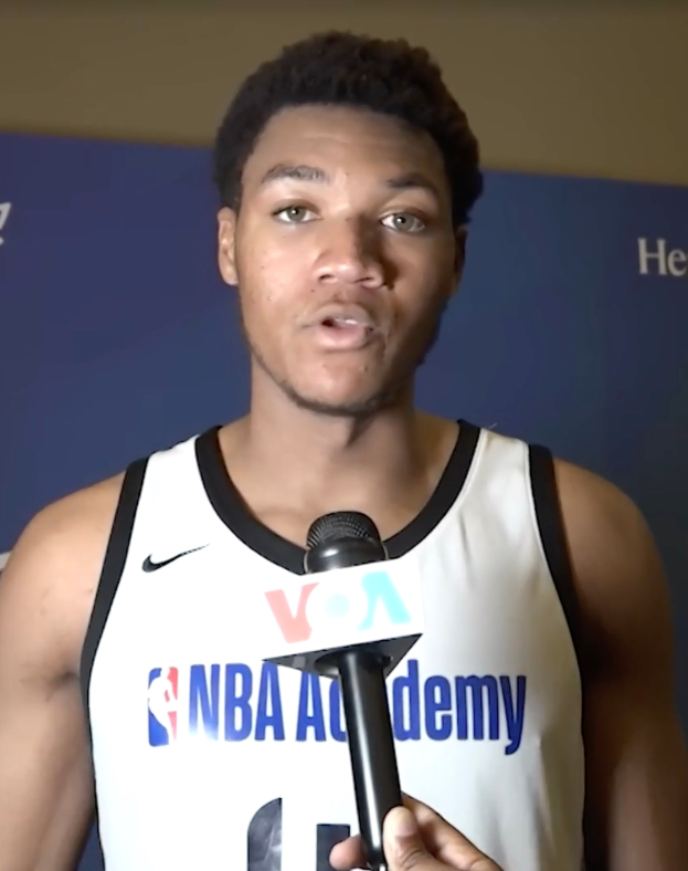 Ein Mann in einem weißen Tanktop hält ein Mikrofon und steht vor einer Tafel mit der Aufschrift "NBA All-Star Game 2019" und einer Wand, wahrscheinlich spricht er mit den Medien über das bevorstehende Spiel.