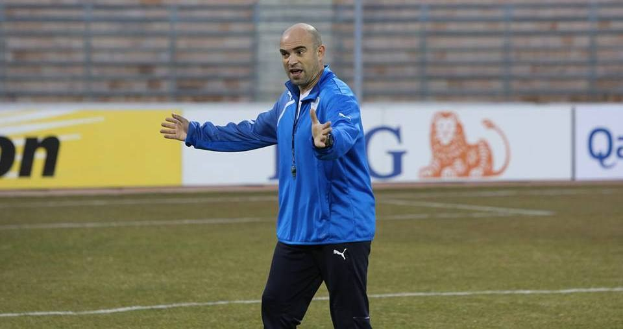 Ein Mann in einer blauen Jacke steht auf einem Fußballfeld während eines Trainings, blickt entschlossen nach vorne, mit sichtbaren Werbetafeln im Hintergrund.