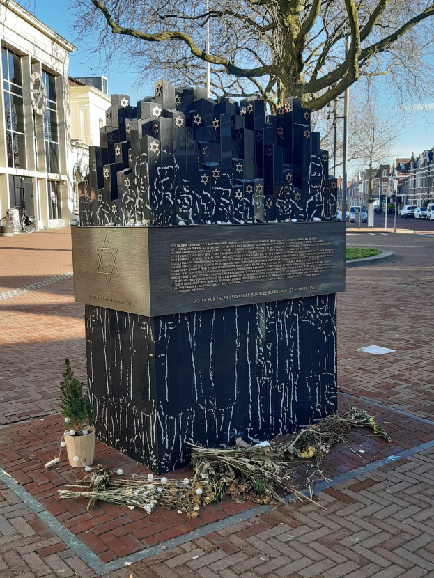 Mahnmal für Holocaust-Opfer in einem Stadtplatz mit Baum, Topfpflanze, verstreuten Blättern und umliegenden städtischen Elementen.