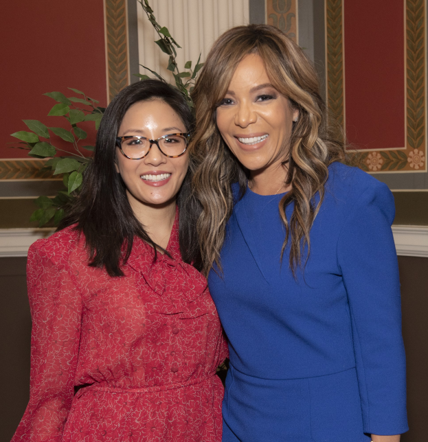 Zwei Frauen, die auf einem Event zusammen lächeln, eine in einem blauen Kleid und die andere in einem roten Kleid mit Brille, vor einer Wand mit einer Pflanze und einem Vorhang im Hintergrund.