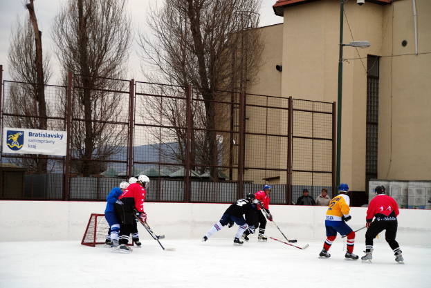Personen beim Eishockeyspielen auf einer Eisfläche mit Gebäuden, Bäumen, einer Straßenlaterne, einem Namensschild und Zäunen im Hintergrund unter einem Himmel.