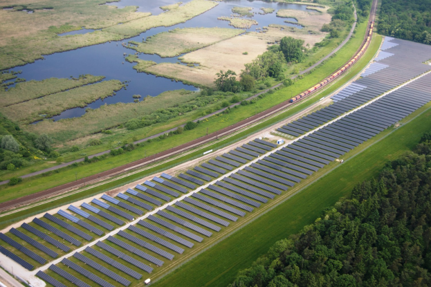 Luftaufnahme eines Solarparks mit Panelen, umgeben von Bäumen, Gras und Wasser, mit einem Zug auf einer nahen Bahnlinie.